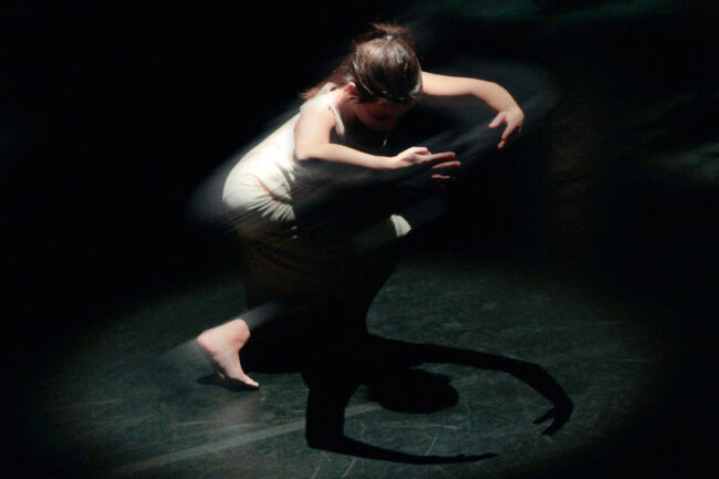 Une femme en robe blanche danse gracieusement dans l'obscurité sur un parquet sombre