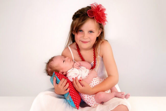 Une petite fille souriante, assise sur un banc dans le studio photo, tient dans ses mains un bébé de trois mois.
