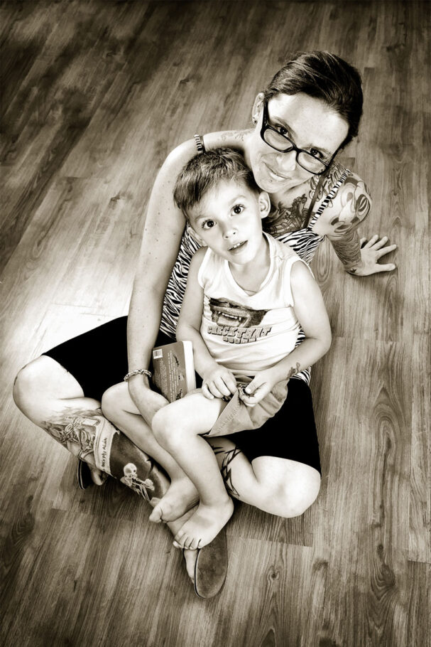 Un petit garçon en short et sa maman tatouée posent assis sur la parquet du studio photo, ambiance noir et blanc sépia.