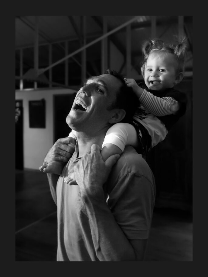 Une petite fille de 3 ans, assises sur les épaules de son père, lui tire les cheveux en souriant.