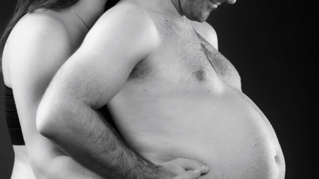 une femme enceinte est derrière son mari torse nu qui gonfle le ventre comme s'il était enceinte