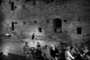 Des gens assis en exterieur avec des murailles derrière eux attendent le début du spectacle, ambiance noir et blanc