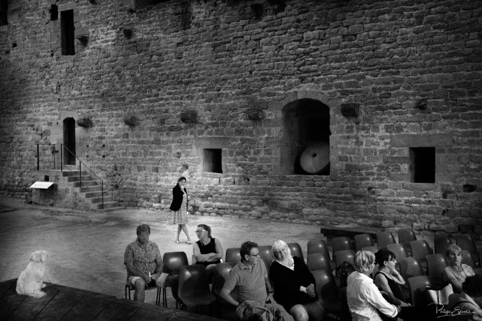 Des gens assis en exterieur avec des murailles derrière eux attendent le début du spectacle, ambiance noir et blanc