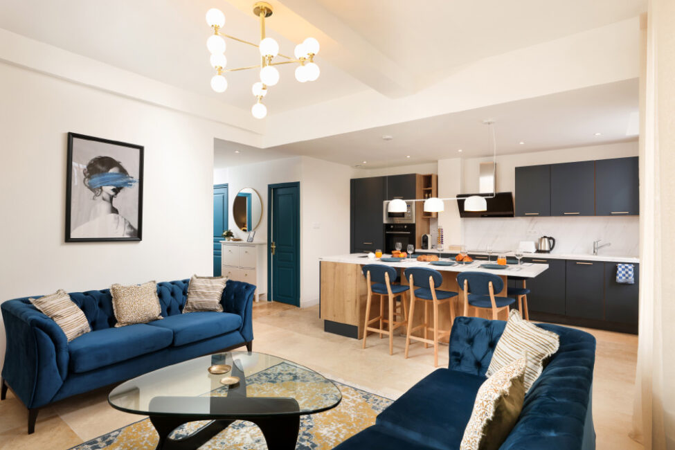 grand salon avec des canapés et des chaises de couleur bleue et en arrière plan une cuisine avec une table dressée