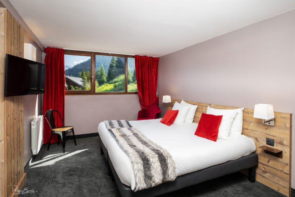 Une chambre d'hôtel avec un lit au drap blanc et coussin rouge, une fenêtre à travers de laquelle on voit un paysage verdoyant et un téléviseur au mur.