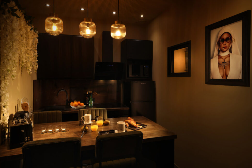 Une cuisine dans la semi obscurité avec une table sur laquelle est dressé un petit déjeuner, verre de jus d'orange, croissant et sur le mur le tableau d'un sœur.