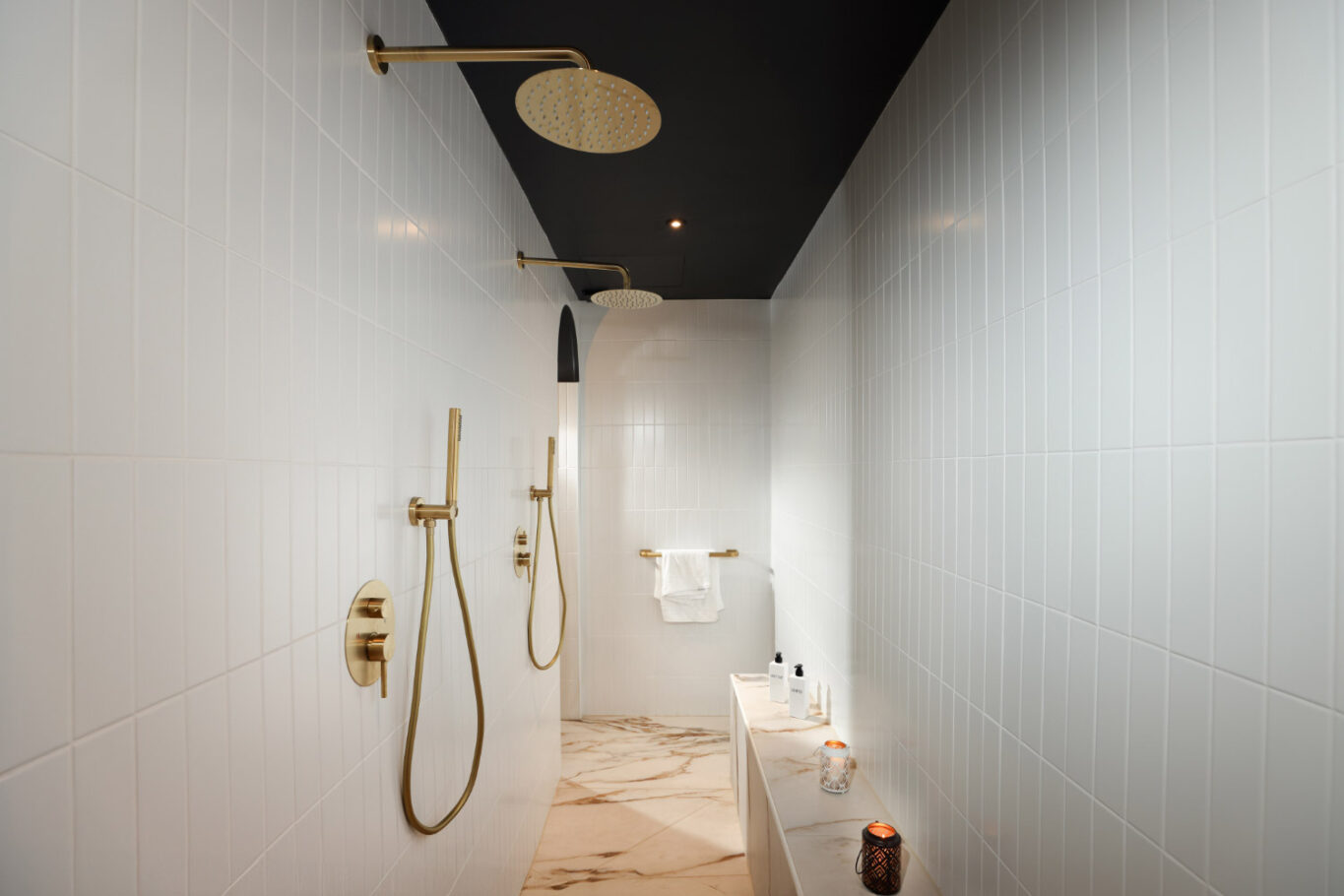 Salle de bain avec un sol en marbre, des murs blancs, deux pommeaux de douche et des photophores allumés.