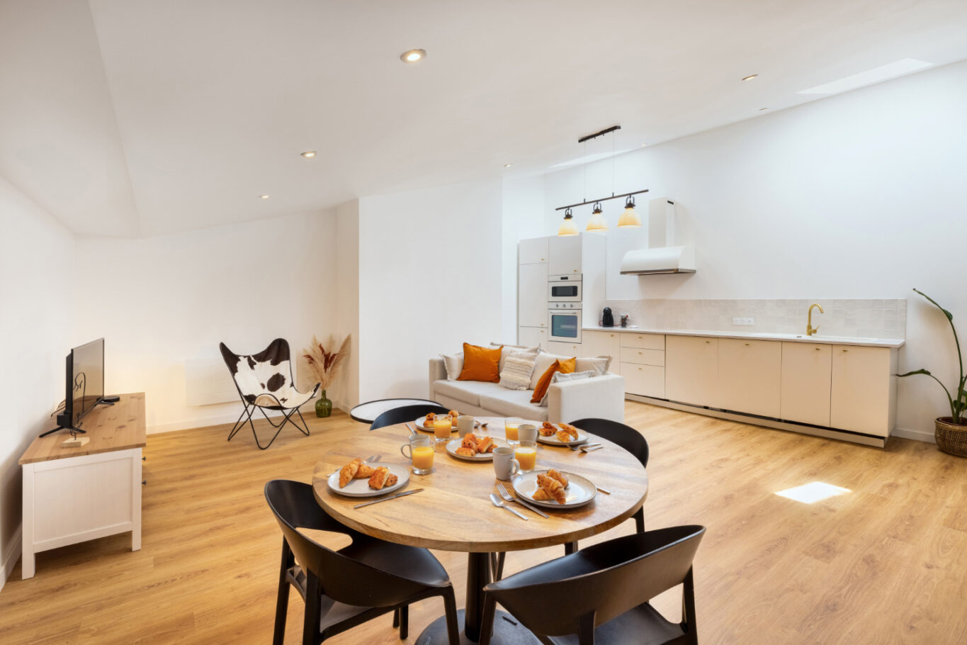 Cuisine et salle à manger blanches avec des sols en bois, une table ronde dressée et un téléviseur sur un meuble.