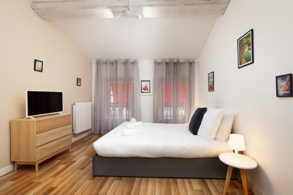 Une chambre avec un sol en bois, un lit aux draps blanc, un téléviseur posé sur un meuble et deux rideaux en arrière plan