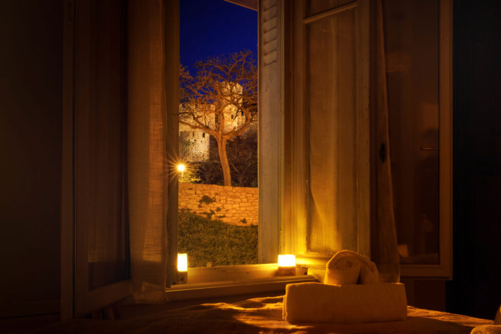 une chambre de nuit, des bougies allumées et une fenêtre ouverte par laquelle on voit un arbre et une partie des rempart la cité de Carcassonne