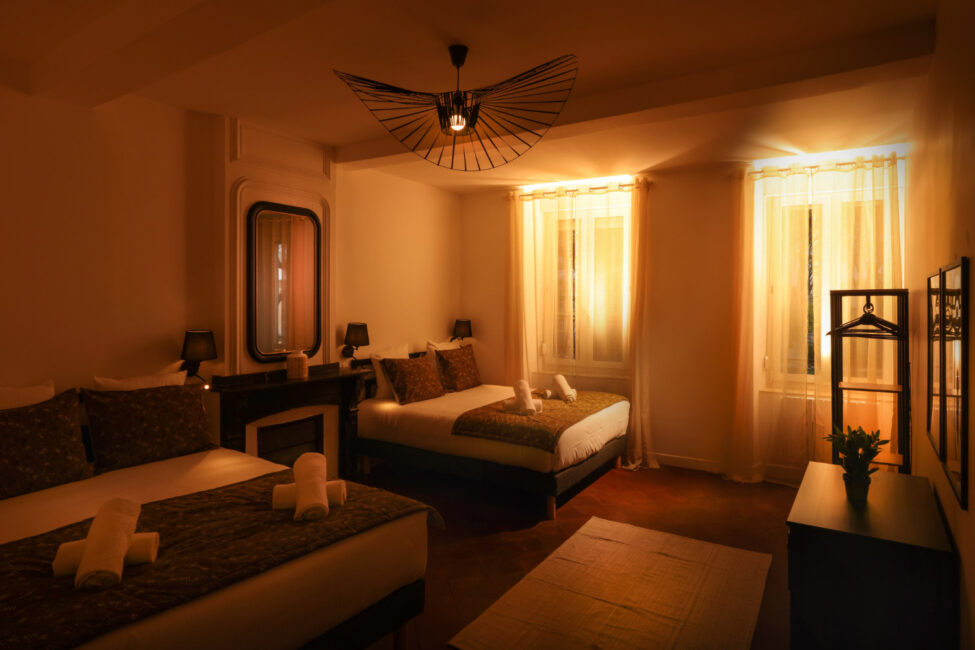 Dans la pénombre, une grande chambre avec des voilages aux fenêtres et deux lits dressés avec des serviettes dessus