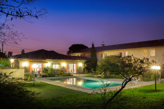 Vue nocturne d'une maison avec piscine, illuminée, évoquant une ambiance tranquille et charmante.