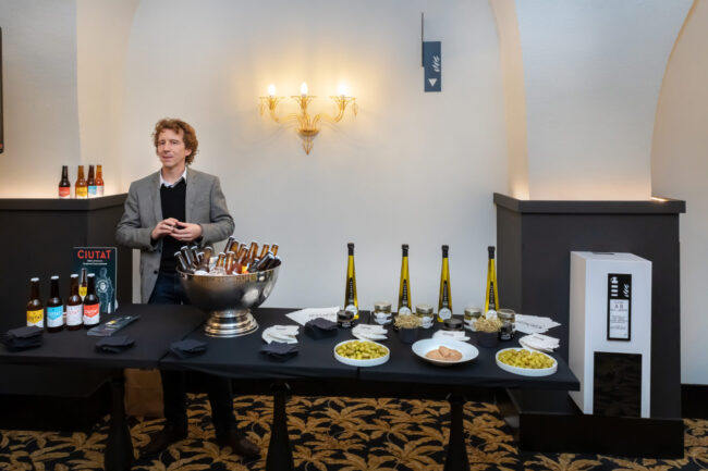 Inauguration de l'hôtel Hilton de Carcassonne 15 Un homme avec devant lui une table sur laquelle il y a des canettes en verre de bière Ciutat et des bouteilles d'huile d'olive