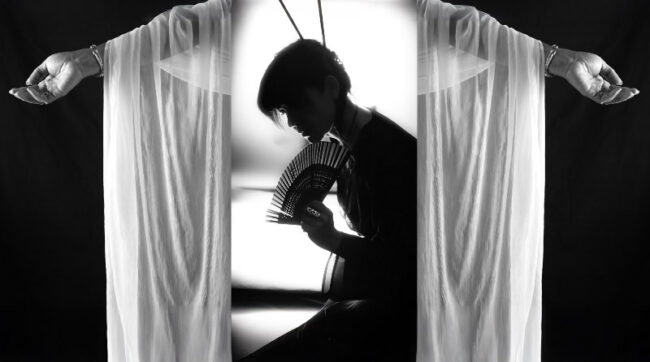 Une femme accroupie en tenue noire tient dans ses mains un éventail, avec deux mains drapées en arrière plan