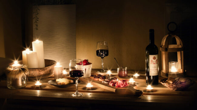 Apéritif servi de nuit sur une table en bois de palette avec une bouteille de vin, deux verres, une planche de charcuterie et des bougies allumées