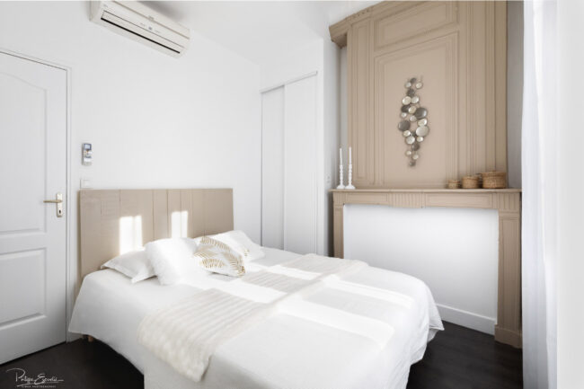 Photos immobilières du mois 2 Chambre à coucher avec un grand lit blanc et une ancienne cheminée obturée en arrière plan.