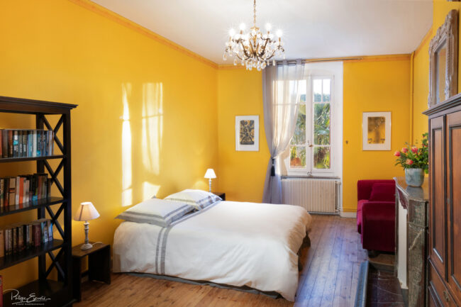 Photos immobilières du mois 5 Chambre de maison de maître avec une étagere, de grands murs jaunes et un lustre ancien au plafond. Une fenêtre en arrière plan devoile de la végétation.