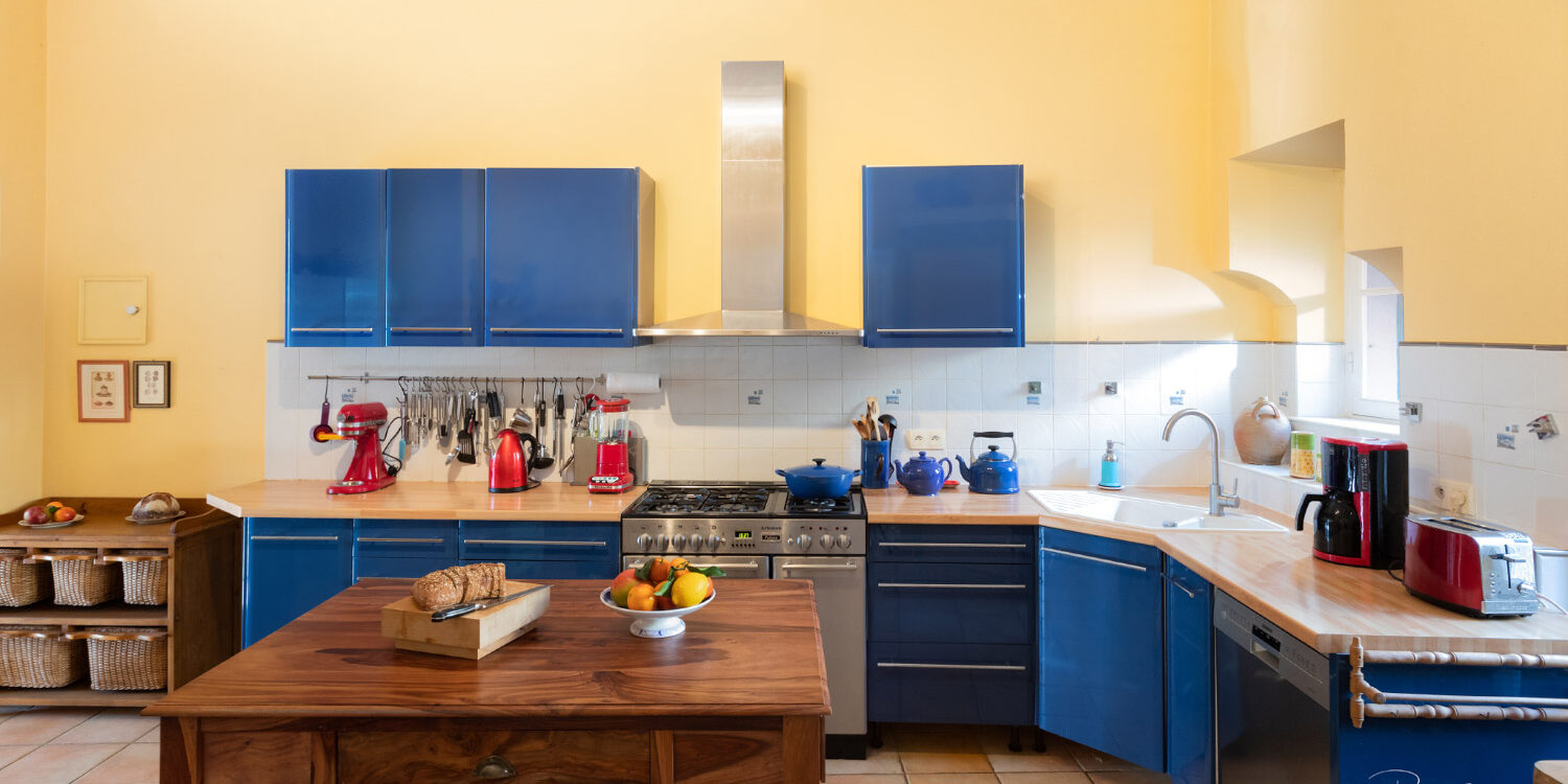Cuisine de maison de maître, avec des placards bleus et une table en bois au premier plan sur laquelle reposent du pain et une corbeille de fruits.