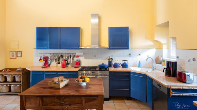 Photos immobilières du mois 6 Cuisine de maison de maître, avec des placards bleus et une table en bois au premier plan sur laquelle reposent du pain et une corbeille de fruits.
