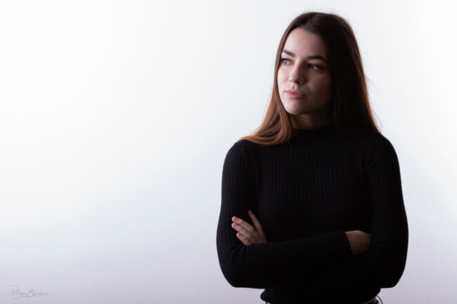 Un jeune fille sur fond blanc, les bras croisés, regarde fixement de trois quart.