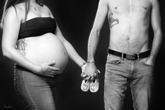 Couple en noir et blanc, dont la femme est enceinte, se tenant la main de face avec une paire de chaussons dans les mains