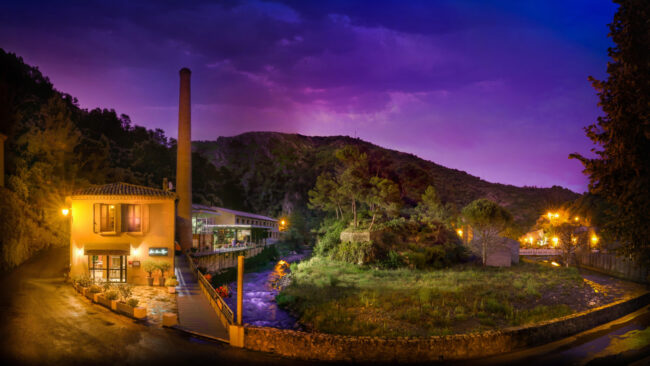Restaurant Le Puits du Trésor, Lastours 1 Ambiance noturne d'éxterieur avec une maison allumée, une riviere et de la végétation
