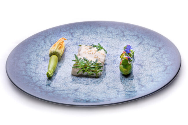 Dans les cuisines du Puits du Trésor, Lastours 6 grande assiette bleue avec dedans une fleur de courgette, un poisson avec une crème blanche dessus et une courgette avec des fleurs