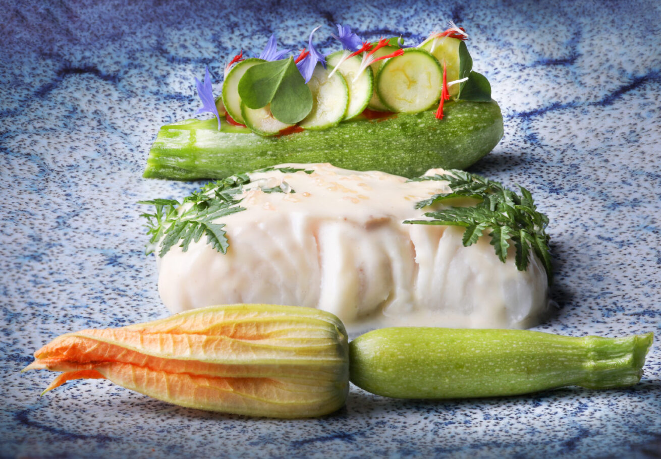 sur une assiette bleue sont posées une fleur de courgette un poisson en sauce blanche et une mini courgette