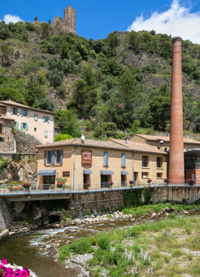 un restaurant avec à ses pieds un ruisseau, de la végétation et au dessus les ruines d'un ancien chateau
