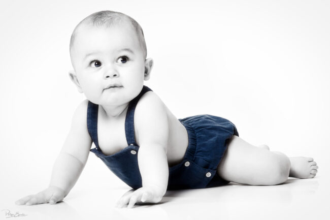 En famille, formule box cadeau ! 2 Un enfant en bas âge en salopette bleue est allongé sur un sol blanc et se redresse sur ses bras.
