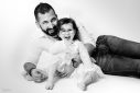 Un papa barbu est allongé sur un fond blanc avec sa fille. Il la tient dans ses bras et les deux sourient.