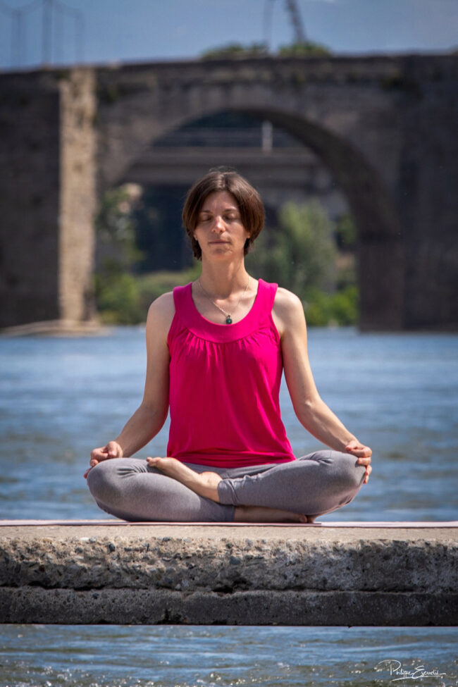 Esprit Tellurique es-tu là ? 3 Une fille assise sur un guet, médite les mains sur les genoux avec une rivière qui passe sous elle et un pont en arrière-plan