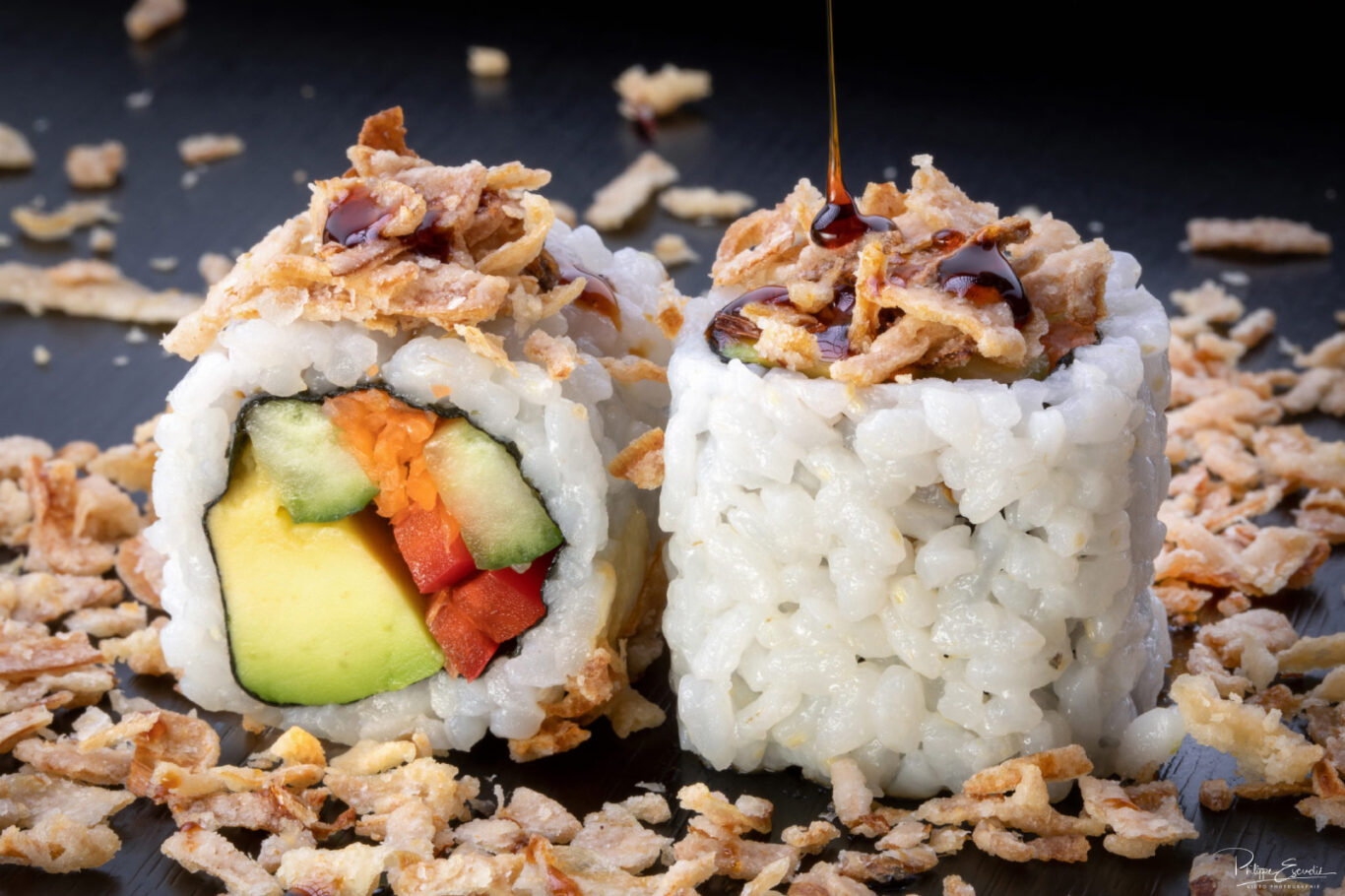 Deux makis végétariens sur une table noire, avec une sauce qui coule dessus et des oignons frits autour.