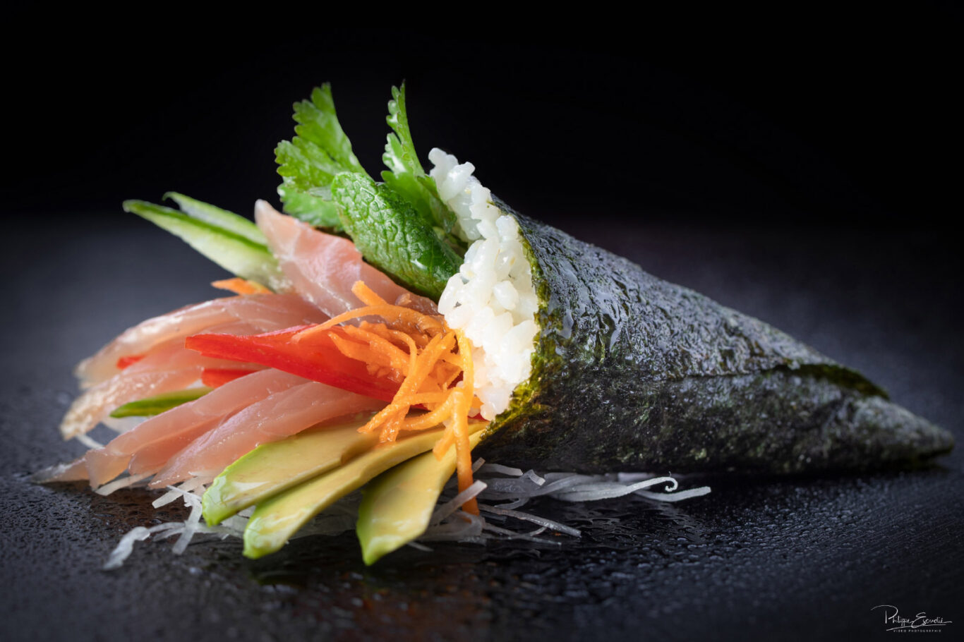 Un temaki en forme de cone, posé sur une table noire, avec à l'intérieur de fines tranches de thon, tomate, carotte et feuille de menthe.