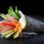 Un temaki en forme de cone, posé sur une table noire, avec à l'intérieur de fines tranches de thon, tomate, carotte et feuille de menthe.