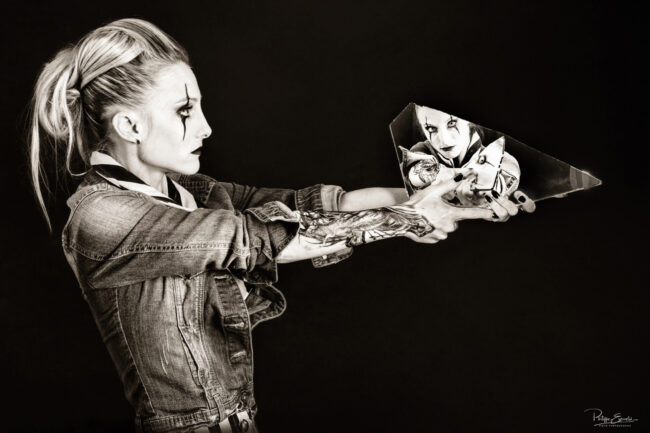 Isa Joker Shooting Studio 3 Une femme blonde de profil, bras tendu, tient dans ses mains un morceau de verre en guise de revolver