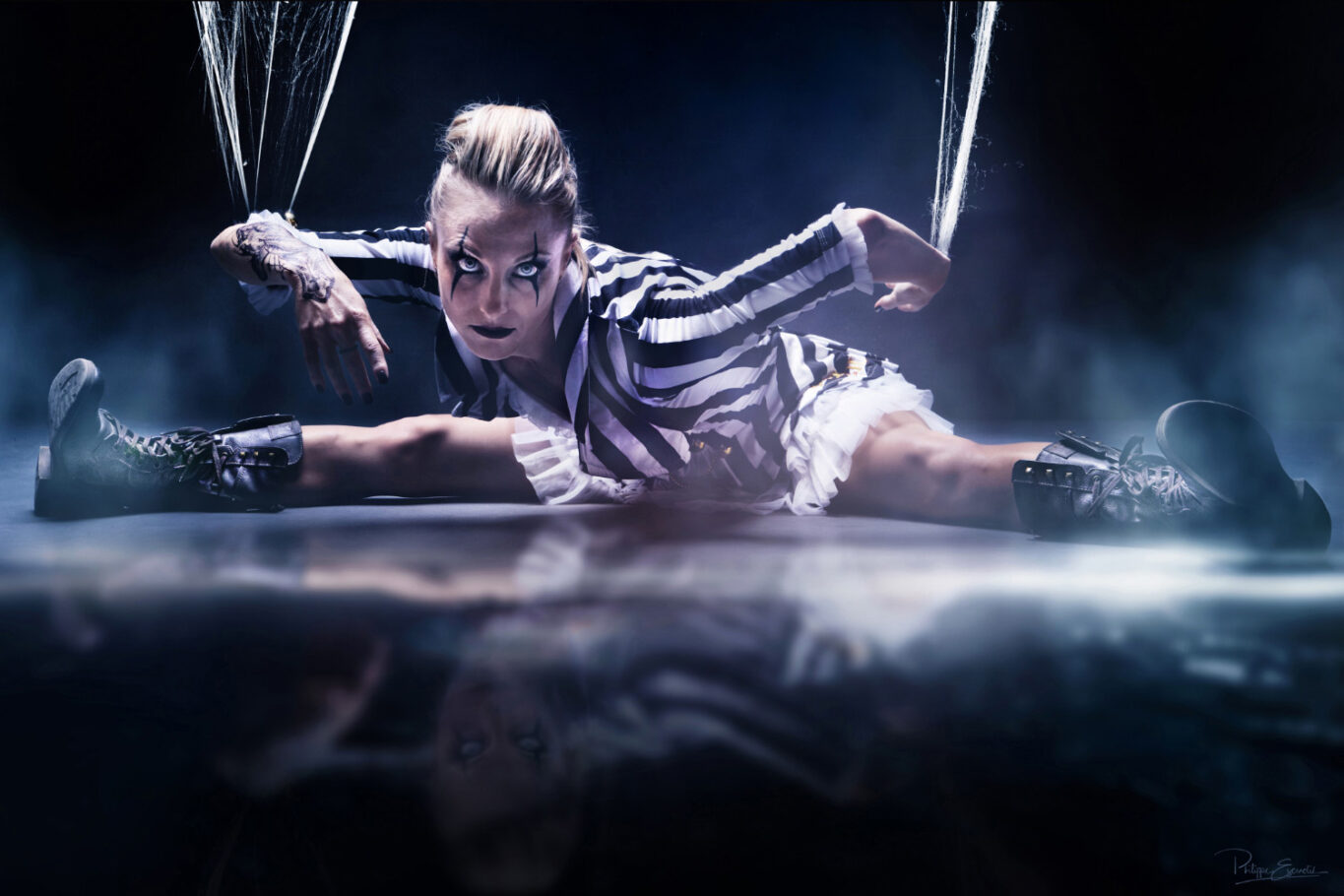 Une femme portant un costume rayé noir et blanc est assise sur le sol en position de grand écart, les bras relevés par des toiles d’araignée comme une marionnette.