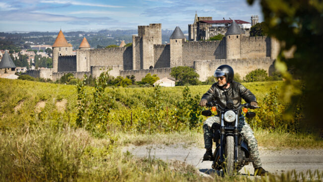 Un motard de face est assis sur une moto avec la Cite de Carcassonne derrière lui