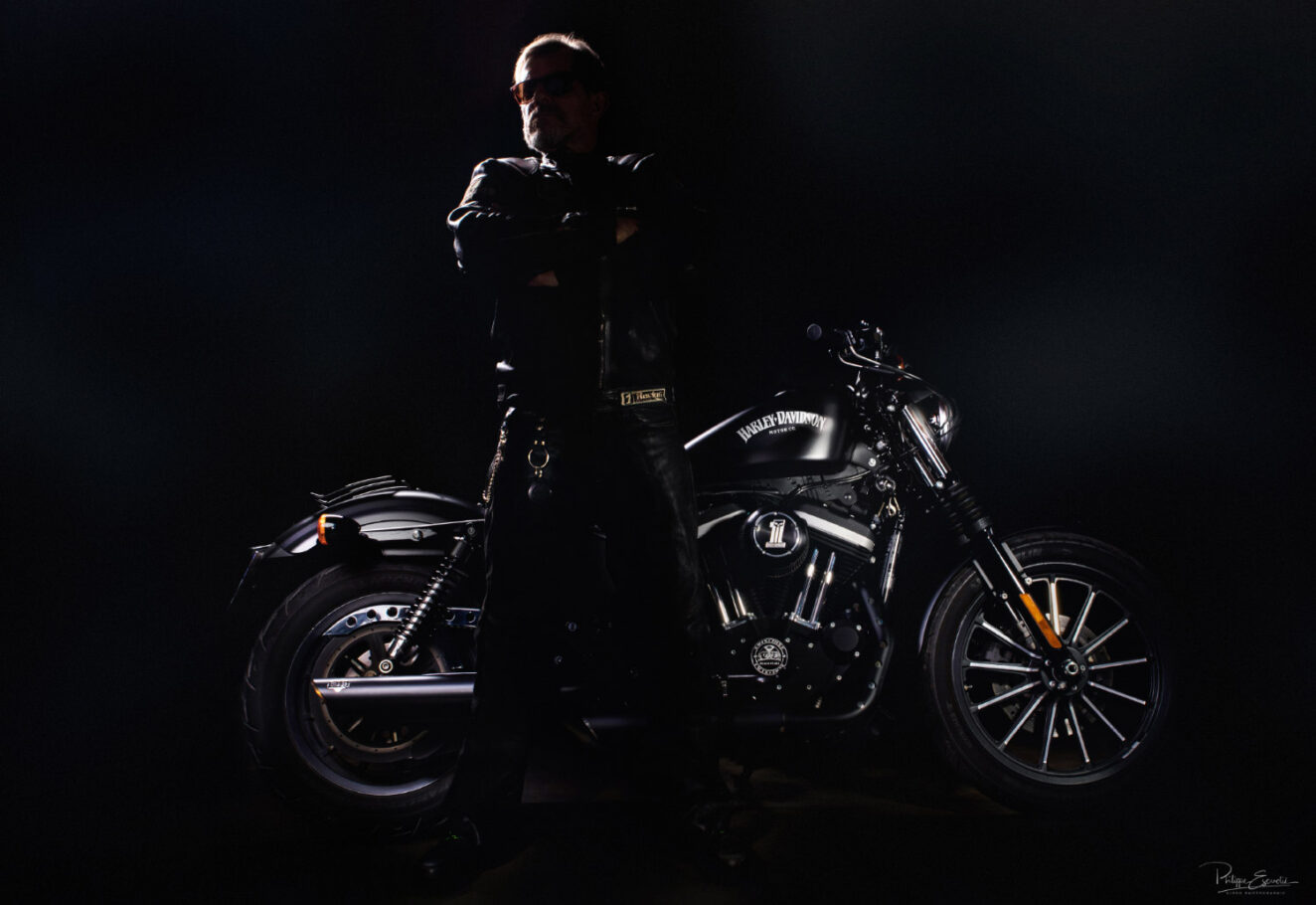 Un motard est debout les bras croisés en contrejour, une moto derrière lui