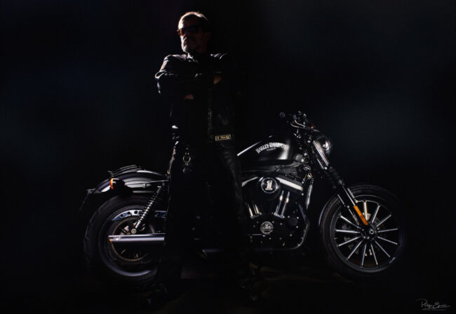 Un motard est debout les bras croisés en contrejour, une moto derrière lui