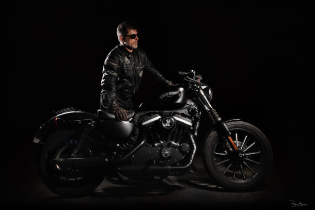 Un motard avec un blouson en cuir est debout derrière sa moto Harley Davidson, une main sur le guidon de la moto, l’autre sur le siège.
