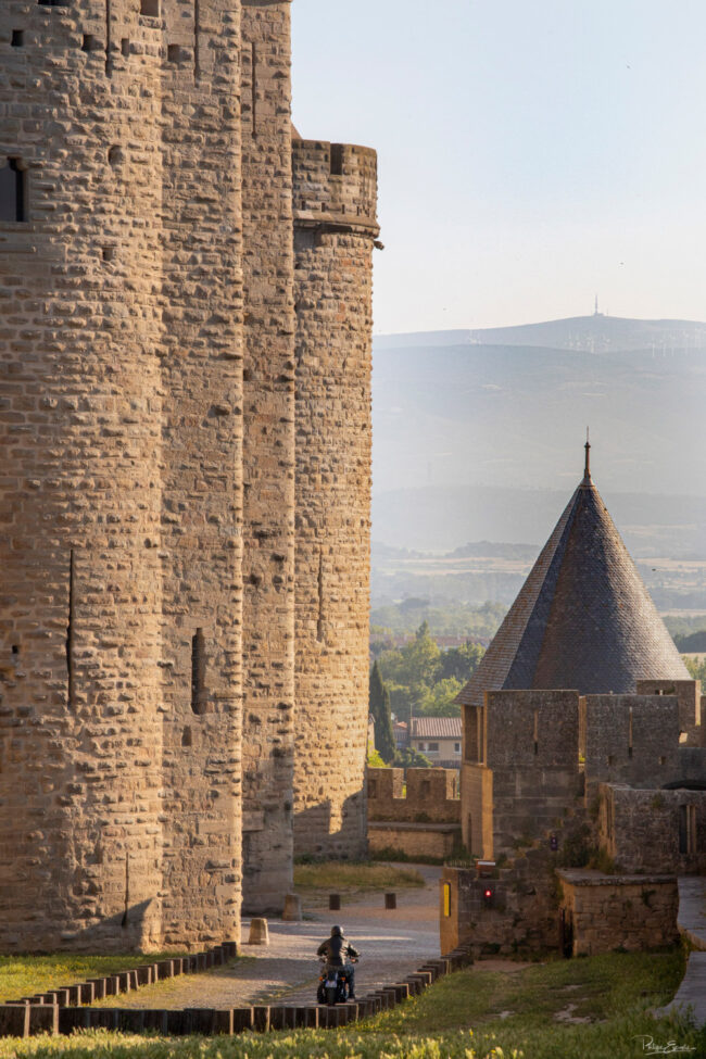 Un motard de profil roule sur son Harley Davidson avec en arrière-plan les murailles floues de la Cité de Carcassonne