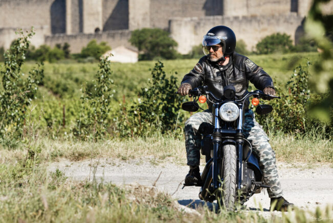 Un motard est à l’arrêt sur sa moto, sur une petite route avec de la végétation en arrière-plan
