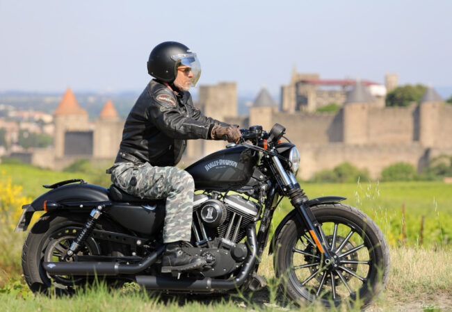 Un motard de profil roule sur son Harley Davidson avec en arrière-plan les murailles floues de la Cité de Carcassonne