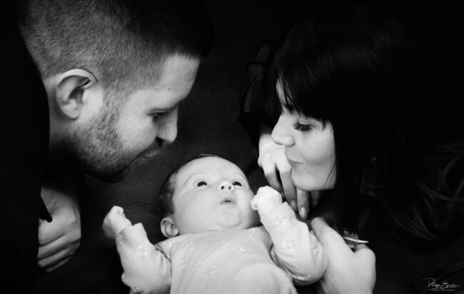 Séance photo nouveau-né et parents 5 Un bébé est allongé sur le dos entouré de ses deux parents qui vont l’embrasser