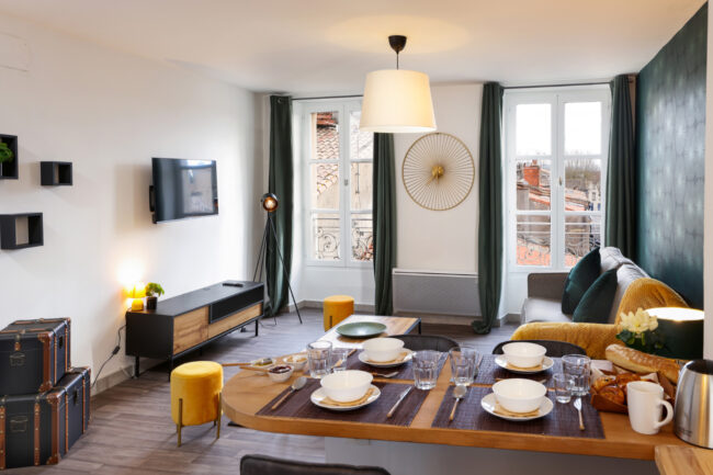 Shooting immobilier, pour maisons d’hôtes, gîtes, hôtels, propriétaires ou conciergerie. 13 Espace cuisine salon avec une table dressée, un canapé et une télé fixée au mur.