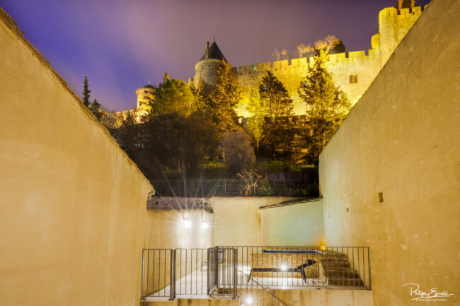 Shooting immobilier, pour maisons d’hôtes, gîtes, hôtels, propriétaires ou conciergerie. 1 Terrasse de nuit aux murs jaunes avec un jacuzzi éclairé et la cité de Carcassonne en arrière-plan.
