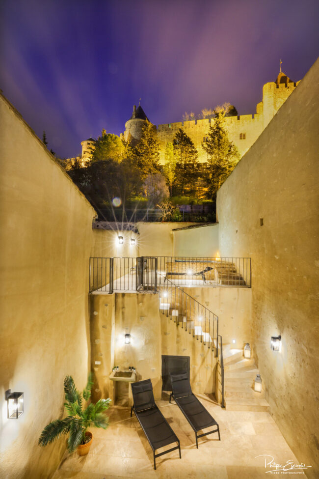 Shooting immobilier, pour maisons d’hôtes, gîtes, hôtels, propriétaires ou conciergerie. 16 Terrasse de nuit avec deux fauteuils relax au sol, un jacuzzi en haut d’un escalier et la cité de Carcassonne en arrière-plan.