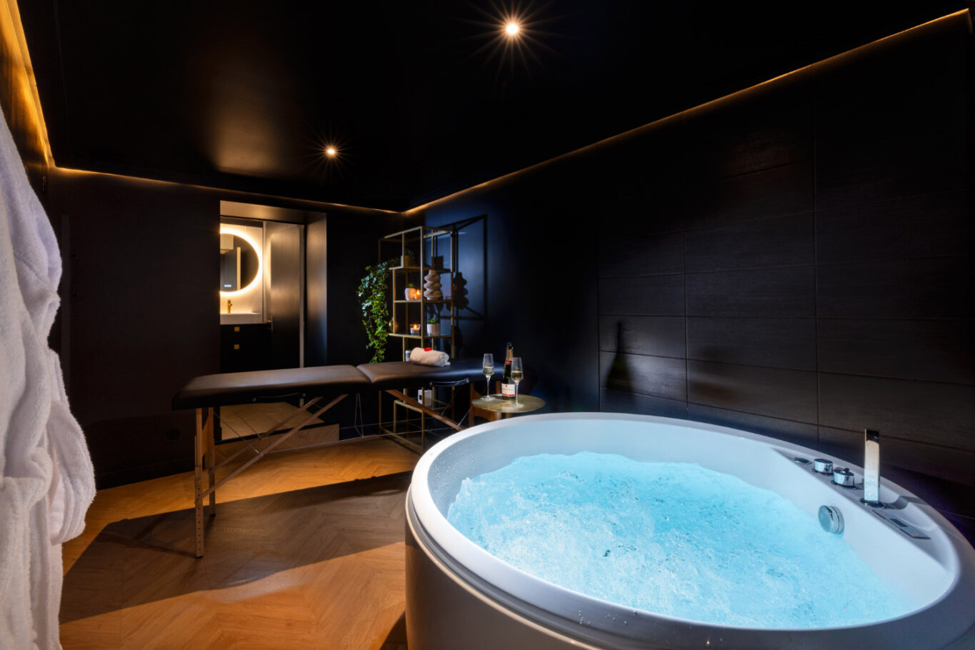 Dans une salle obscure et intime, une grande baignoire balnéo à remous, une table à masser et une tablette avec une bouteille de champagne et deux verres