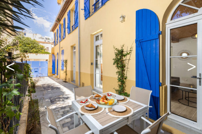 terrasse extérieure avec un petit déjeuner sur une table et une façade jaune aux volets bleu en arrière plan